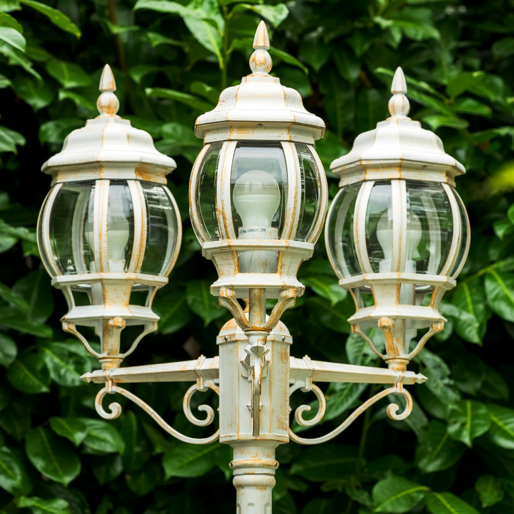 Ein weißer Laternenpfahl mit drei Lampen steht vor grünem Laub.