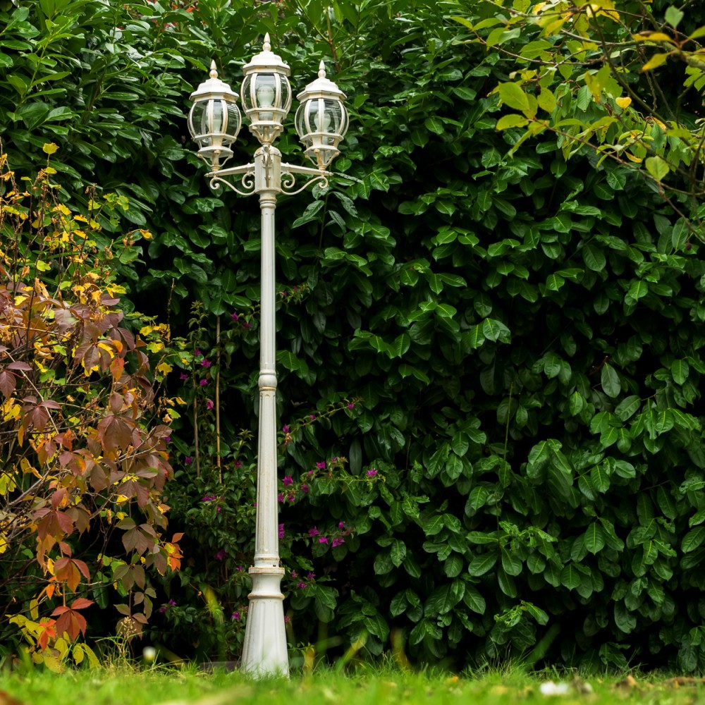 Eine weiße dreiköpfige Laterne steht in einem Garten mit üppigem Grün und Herbstlaub.
