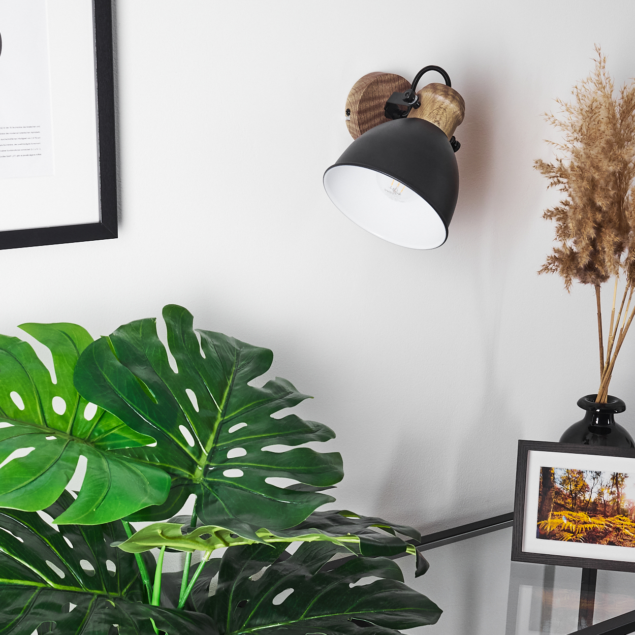 Eine schwarze Wandleuchte mit Holzakzenten hängt an einer weißen Wand, neben einer Monstera-Pflanze.