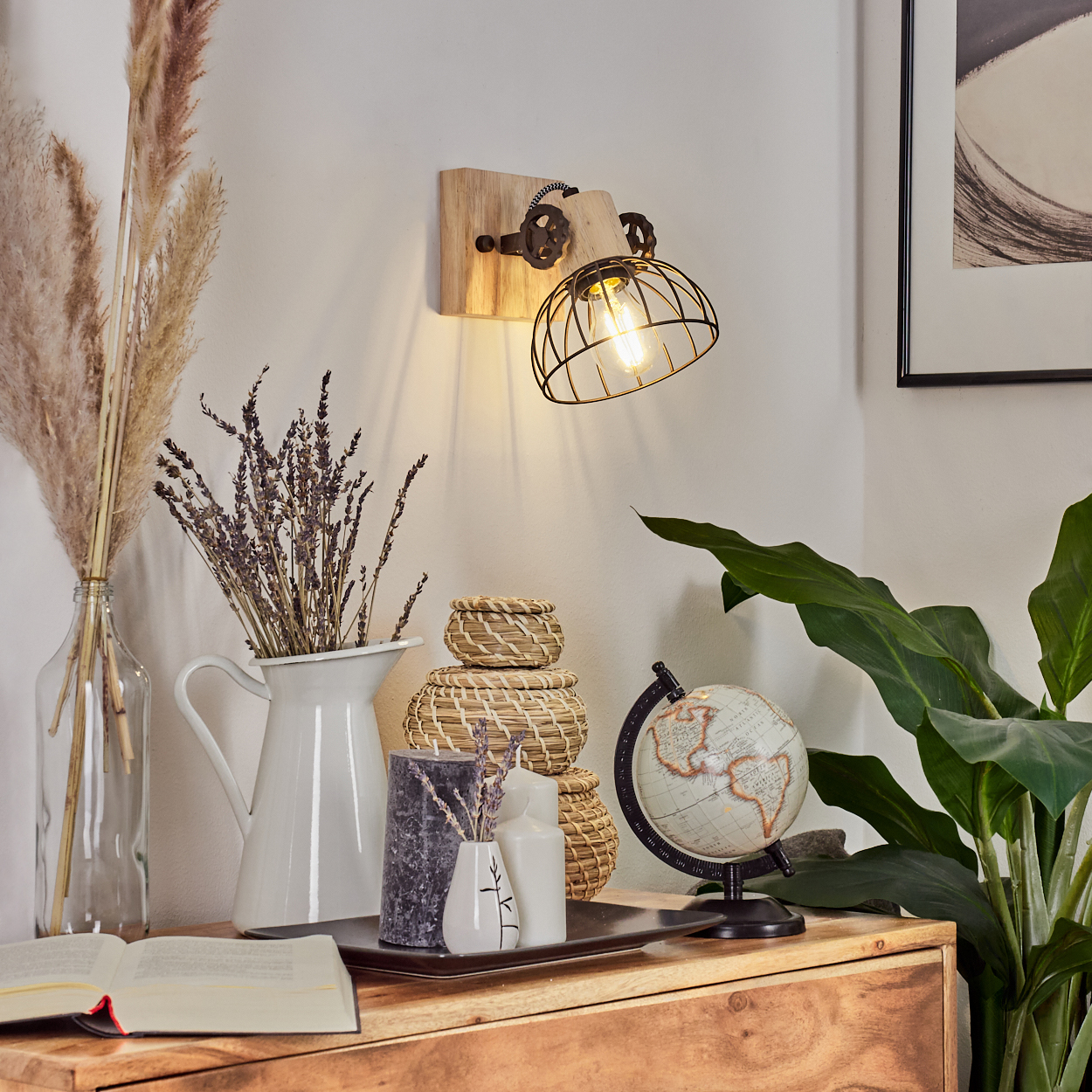 Eine rustikale Wandleuchte beleuchtet eine Holzkommode mit Deko: Globus, Pflanzen, Kerzen, Buch.
