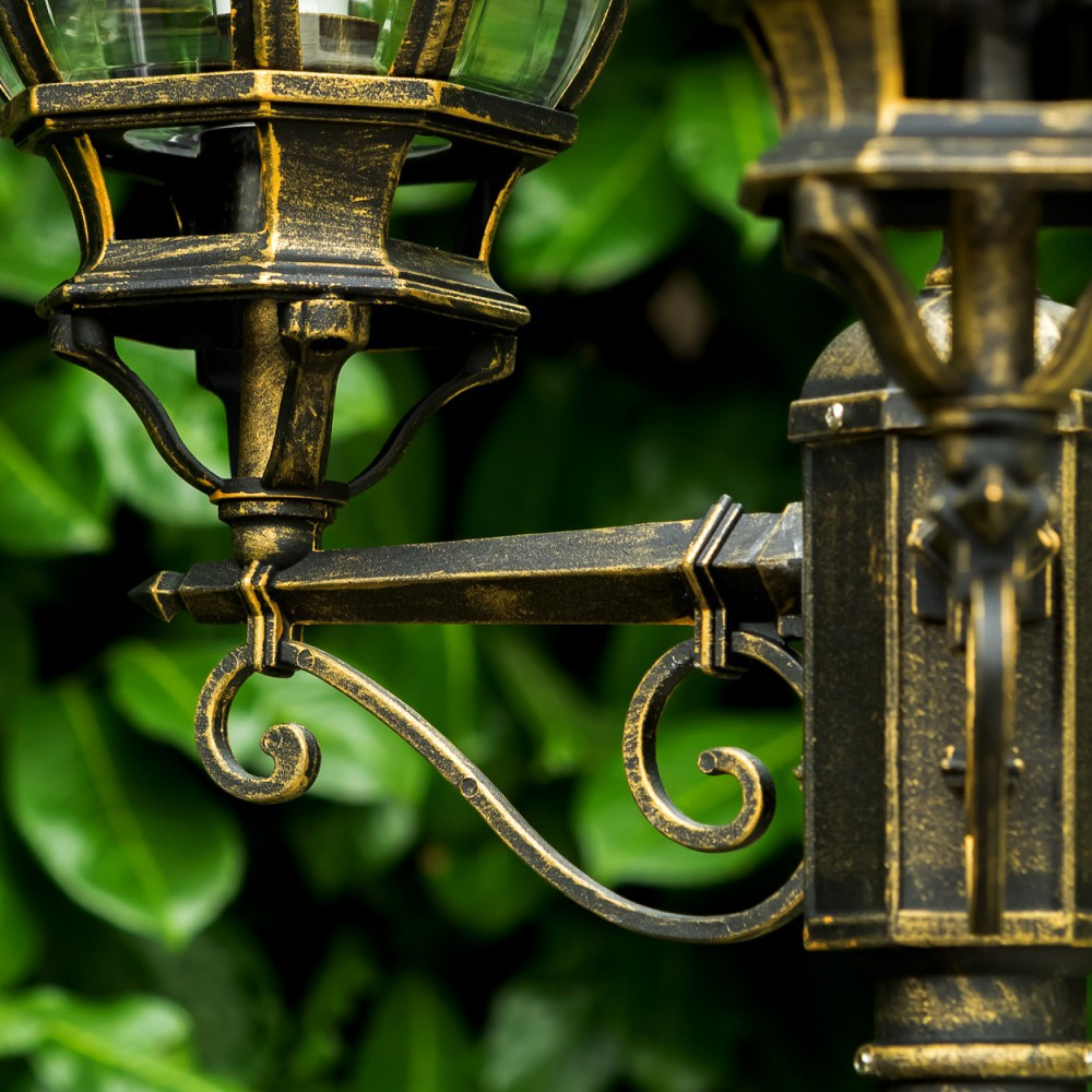 Detail einer antiken Lampe aus Messing mit kunstvollen Schnörkeln vor grünem Laub.