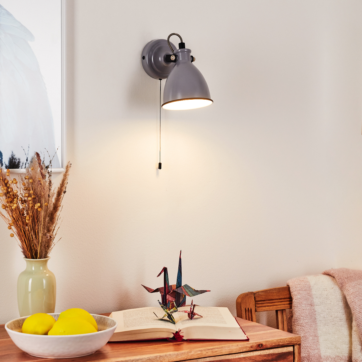 Graue Wandlampe über einem offenen Buch mit Origami-Kranichen, einer Vase mit Trockenblumen und Zitronen.