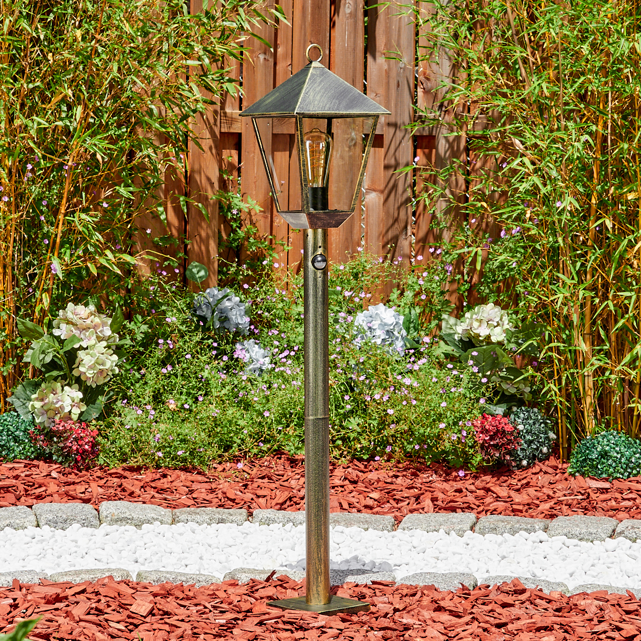 Gartenlaterne mit Glühbirne vor Bambus und Blumen, mit Rindenmulch und weißen Steinen.