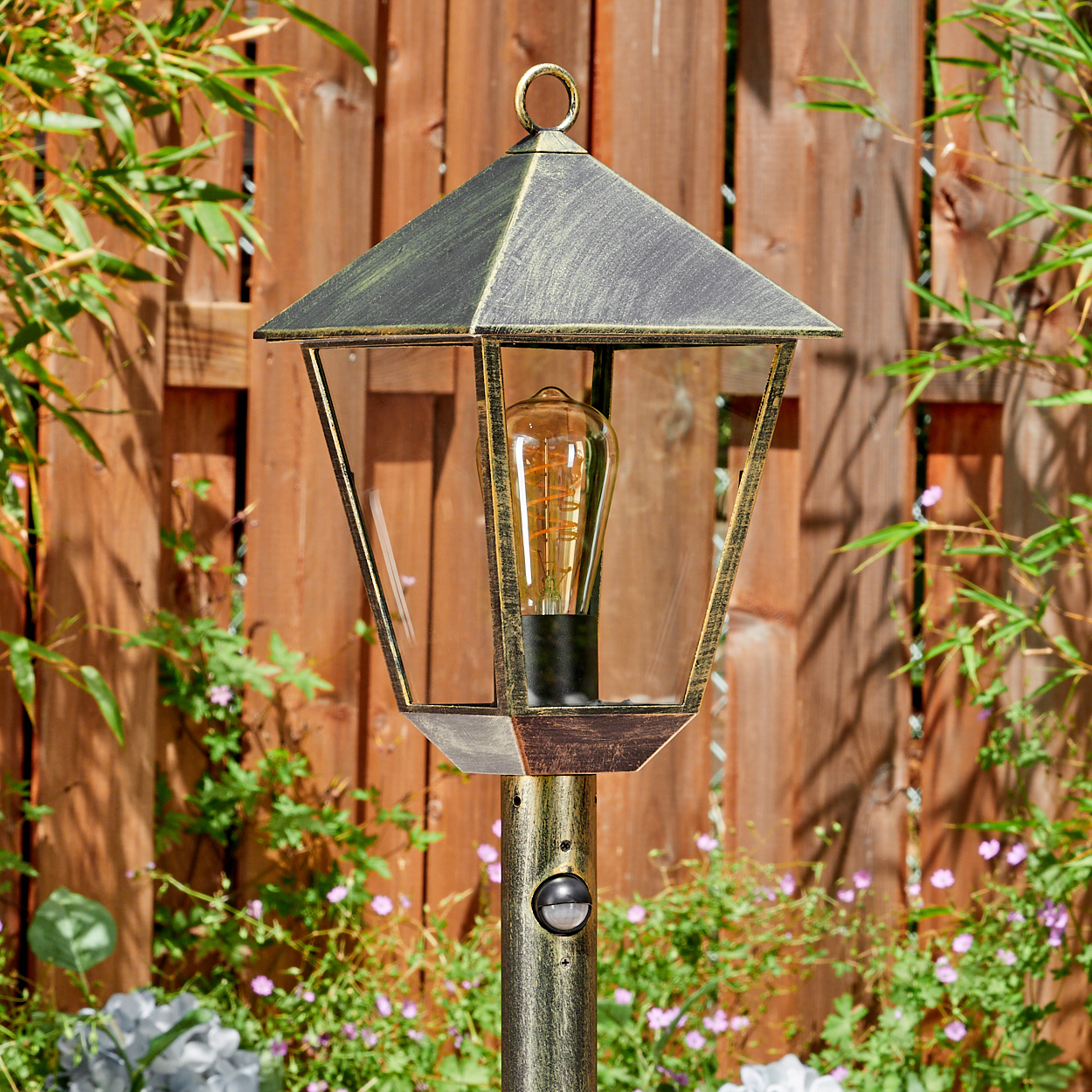 Eine goldene Laterne mit Glühbirne steht im Garten vor einem Holzzaun.