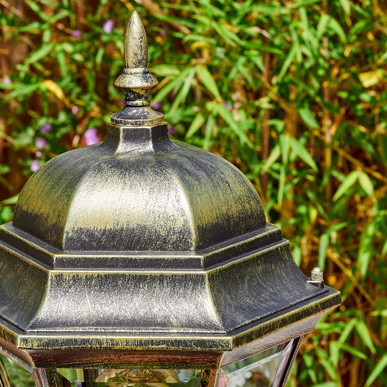 Detail eines schwarzen und goldenen Laternenkopfes vor grünem Laub.