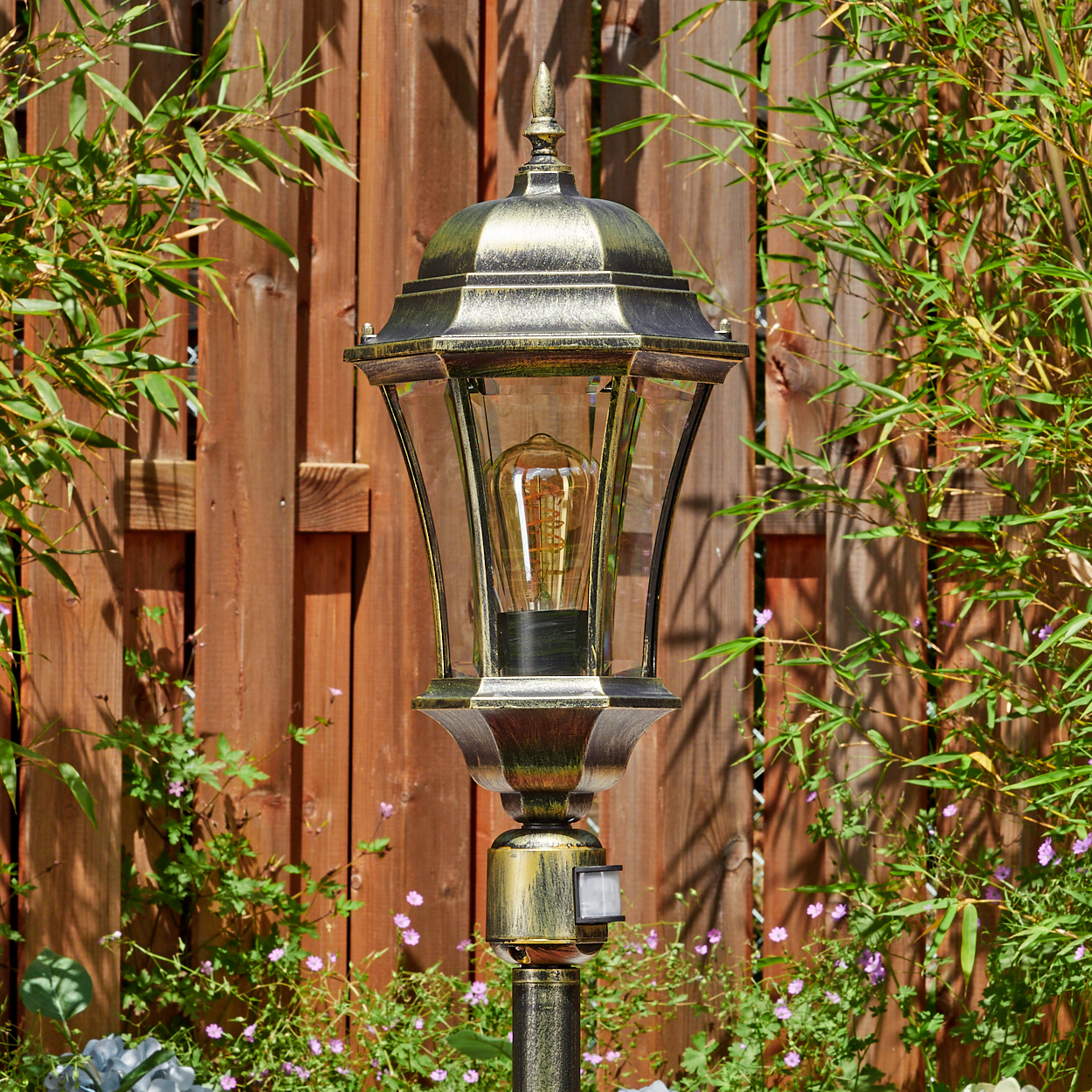 Eine goldene Lampe mit Glühbirne steht vor einem Holzzaun und Bambus.