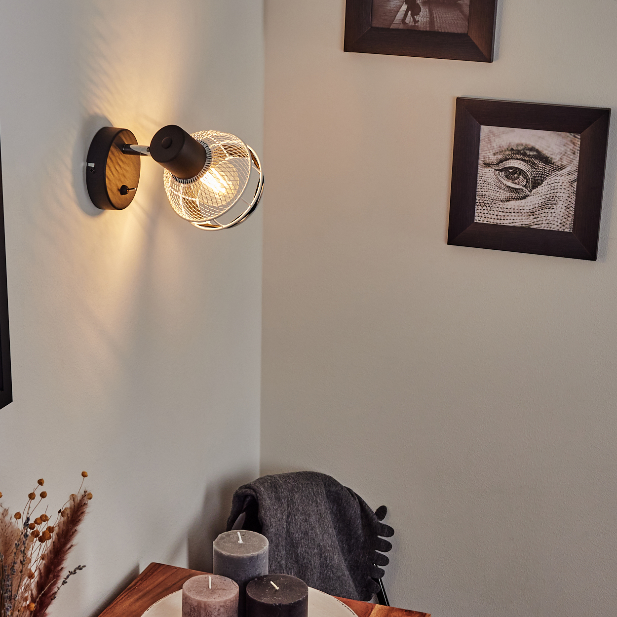 Eine Wandleuchte mit Metallkäfig wirft warmes Licht auf eine Wand mit Bildern.