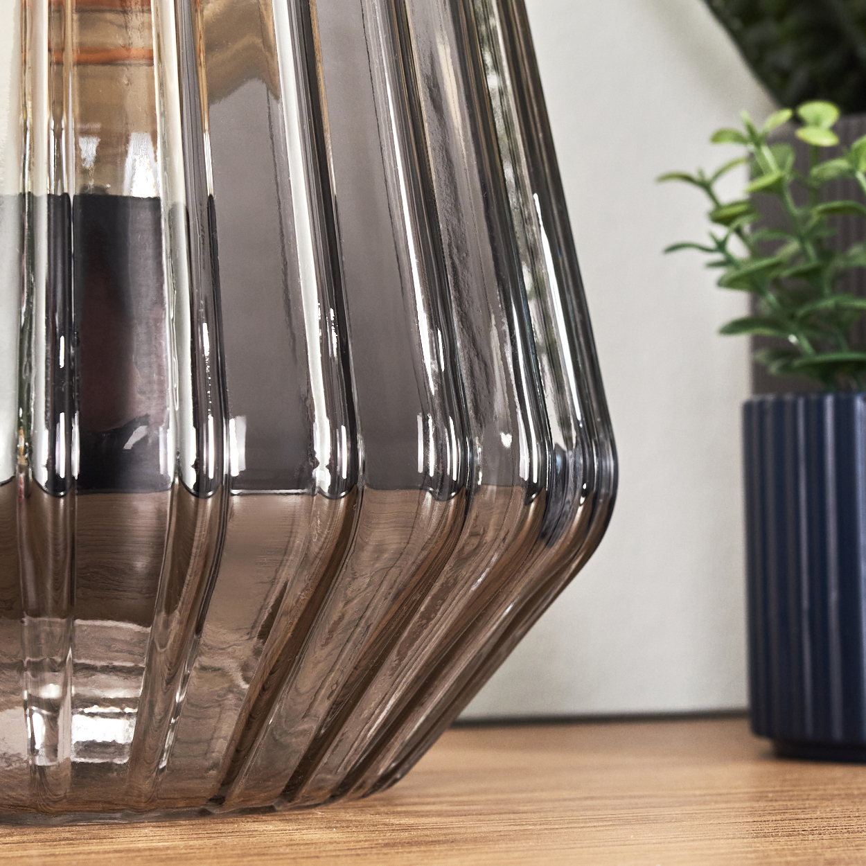 Graue gerippte Vase auf einem Holztisch, daneben eine Topfpflanze.