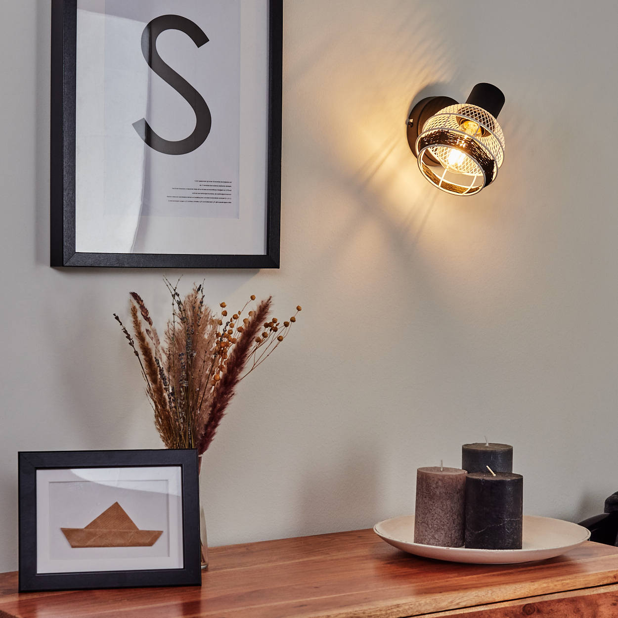 Ein Wandspot beleuchtet eine Wand mit einem gerahmten Bild und Trockenblumen.