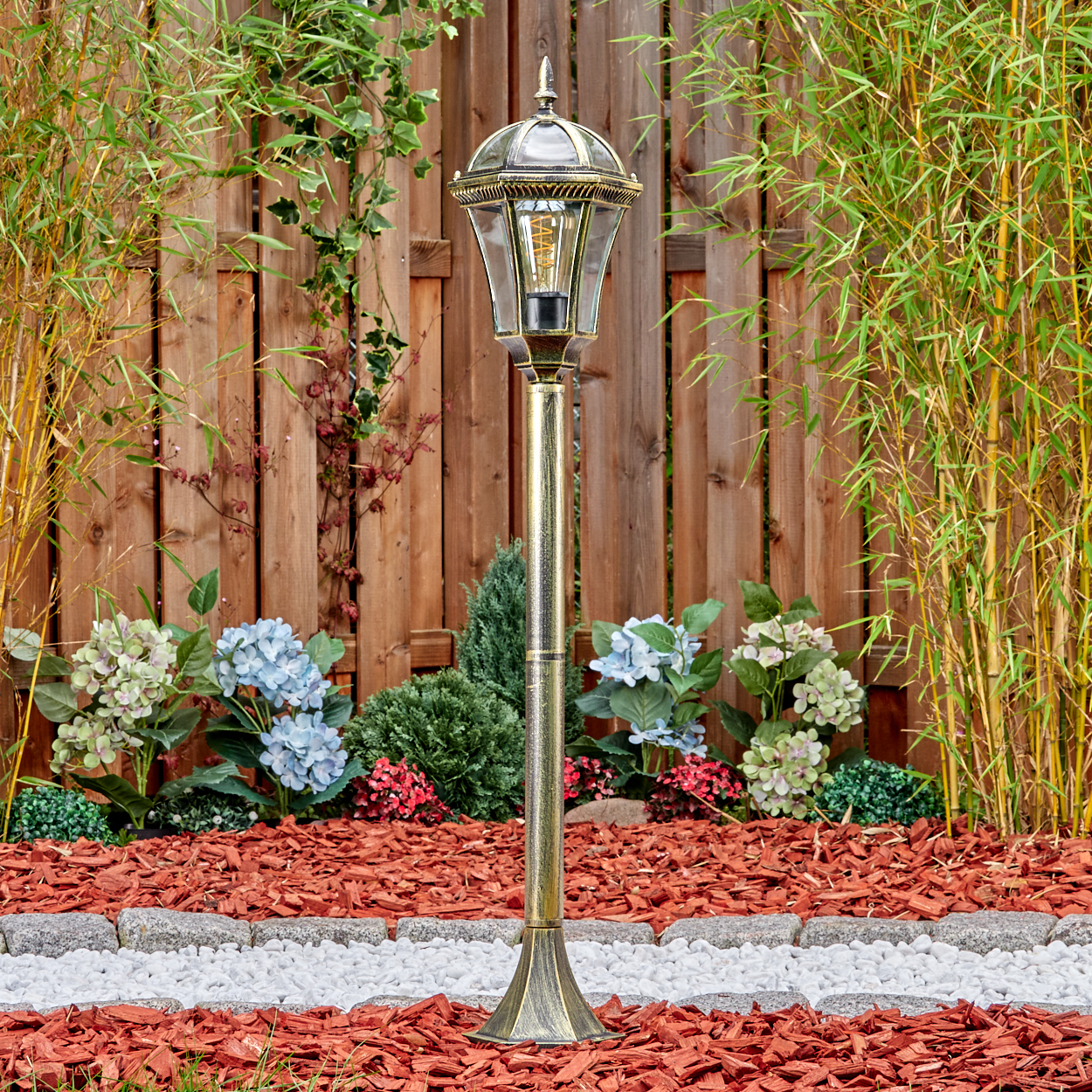 Ein goldener Gartenpfosten mit Glaslaterne steht vor einem Holzzaun und Bambus.