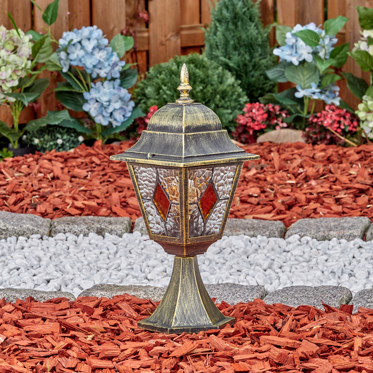 Eine bronzene Gartenlaterne mit Glasfenstern steht auf rotem Mulch vor einem Holzzaun.