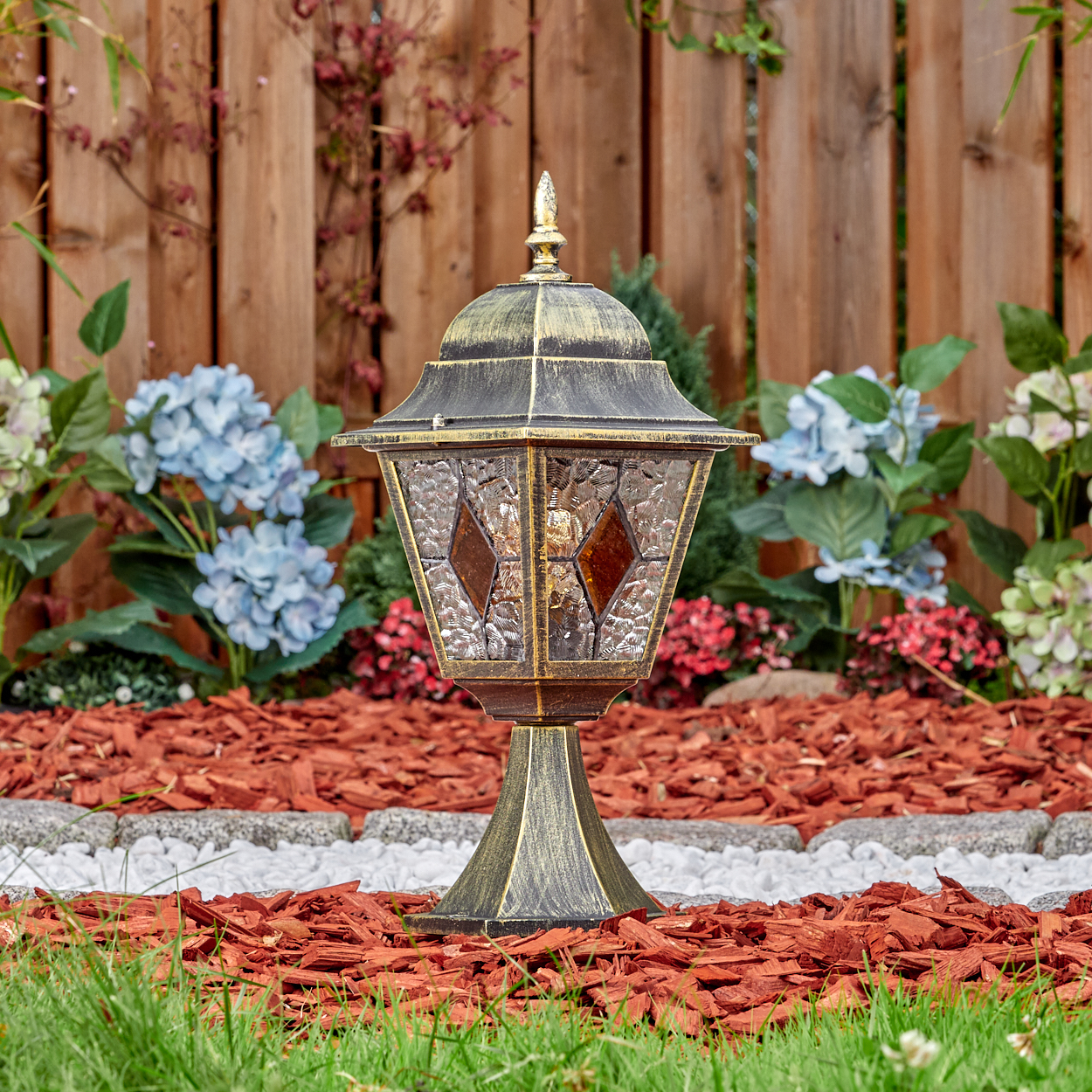 Eine dekorative Gartenlaterne aus Metall mit Glasfenstern steht im Vordergrund, umgeben von Rindenmulch.