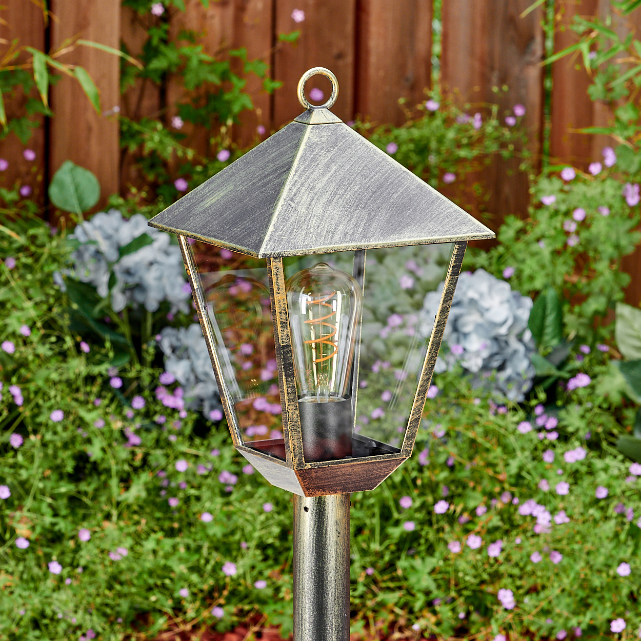 Eine bronzefarbene Gartenlaterne mit Glühbirne steht vor einem Holzzaun und Blumen.