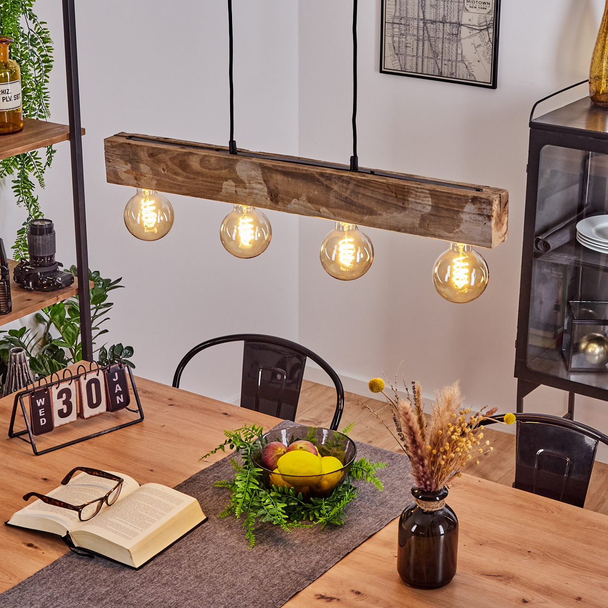 Holzpendelleuchte mit vier Glühbirnen über Holztisch mit Buch, Brille, Obstschale und Vase.