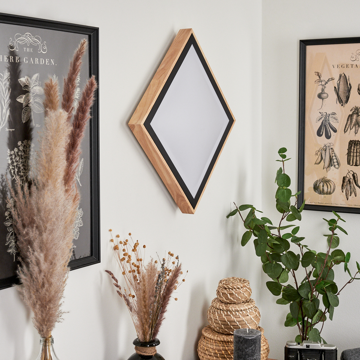 Ein rautenförmiger Spiegel mit Holzrahmen hängt an der Wand neben Bildern.