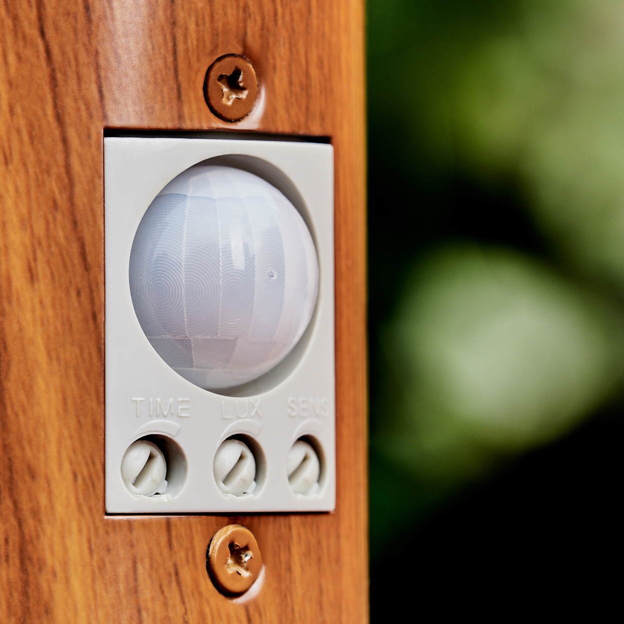 Ein Bewegungsmelder mit weißer Kuppel auf Holz, mit Reglern für Zeit, Lux und Sens.