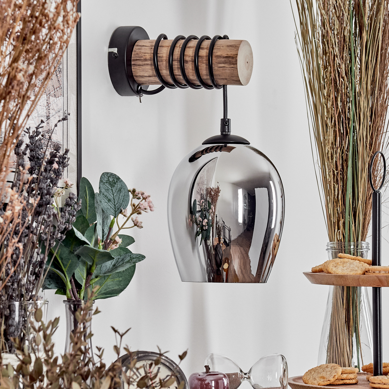 Eine silberne Pendelleuchte mit Holzarm und Metallspiralen hängt an einer weißen Wand.