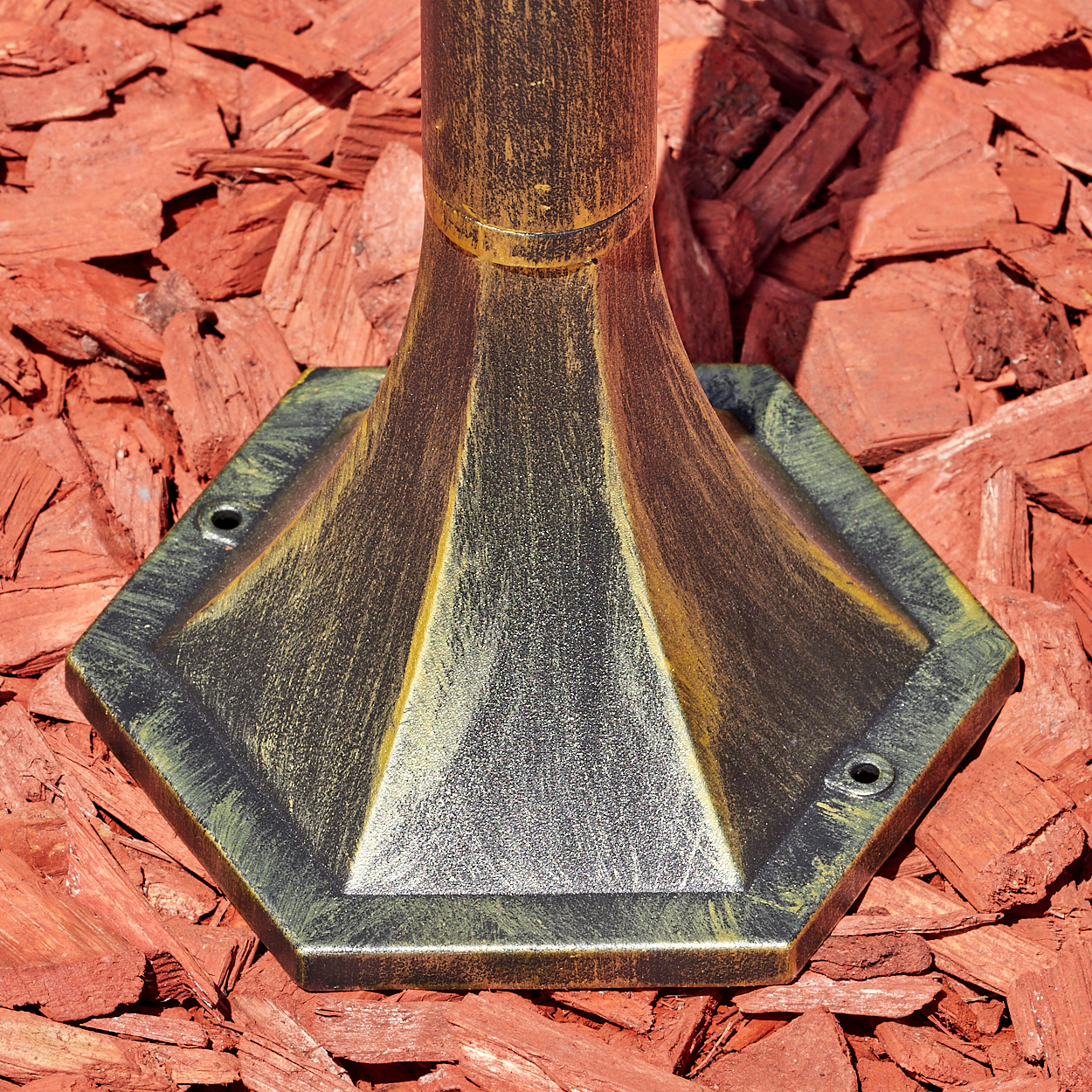 Ein goldfarbener Lampenfuß mit sechseckiger Basis auf rotem Rindenmulch.