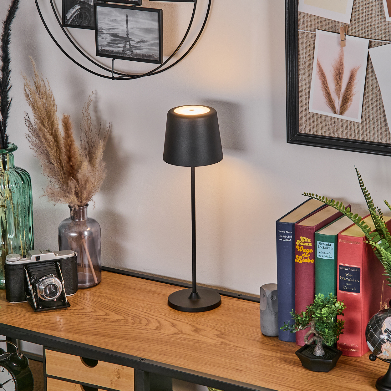 Ein schwarzer Nachttischlampenschirm leuchtet auf einem Holztisch neben Büchern und Pflanzen.