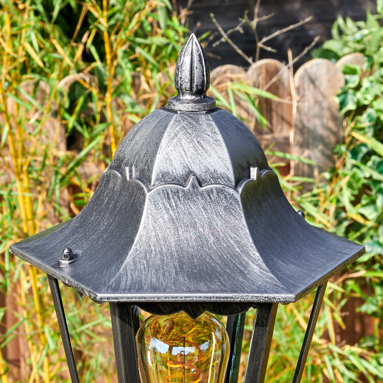Eine silberne Gartenlaterne mit Glühbirne vor Bambus und Zaun.