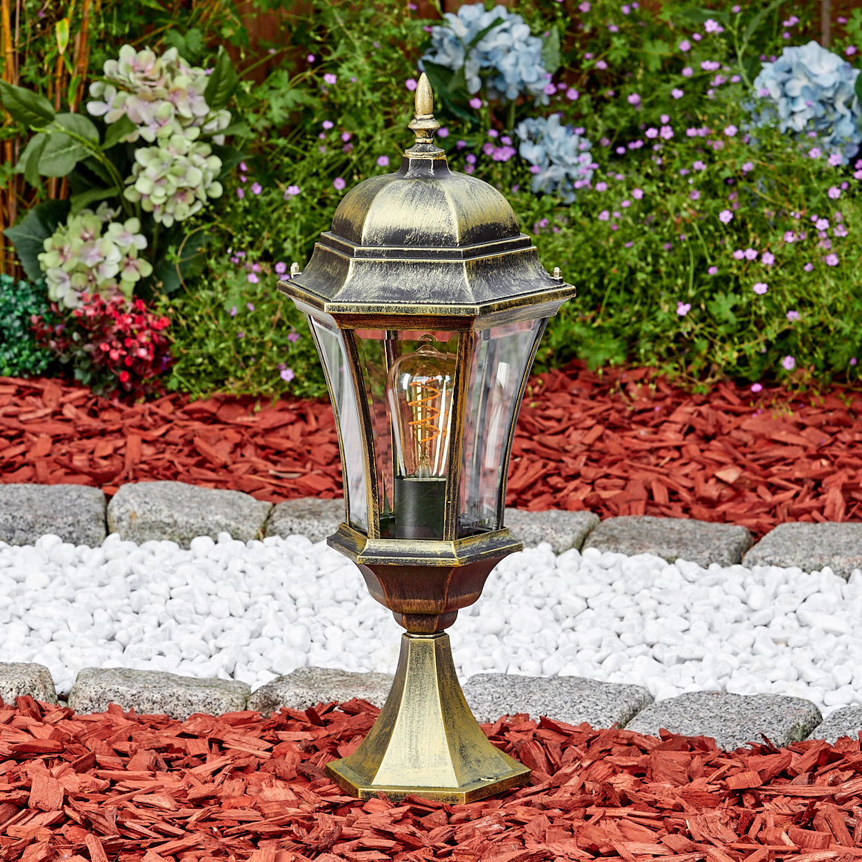 Ein goldener Außenlaternenfuß mit Glühbirne steht im Garten.