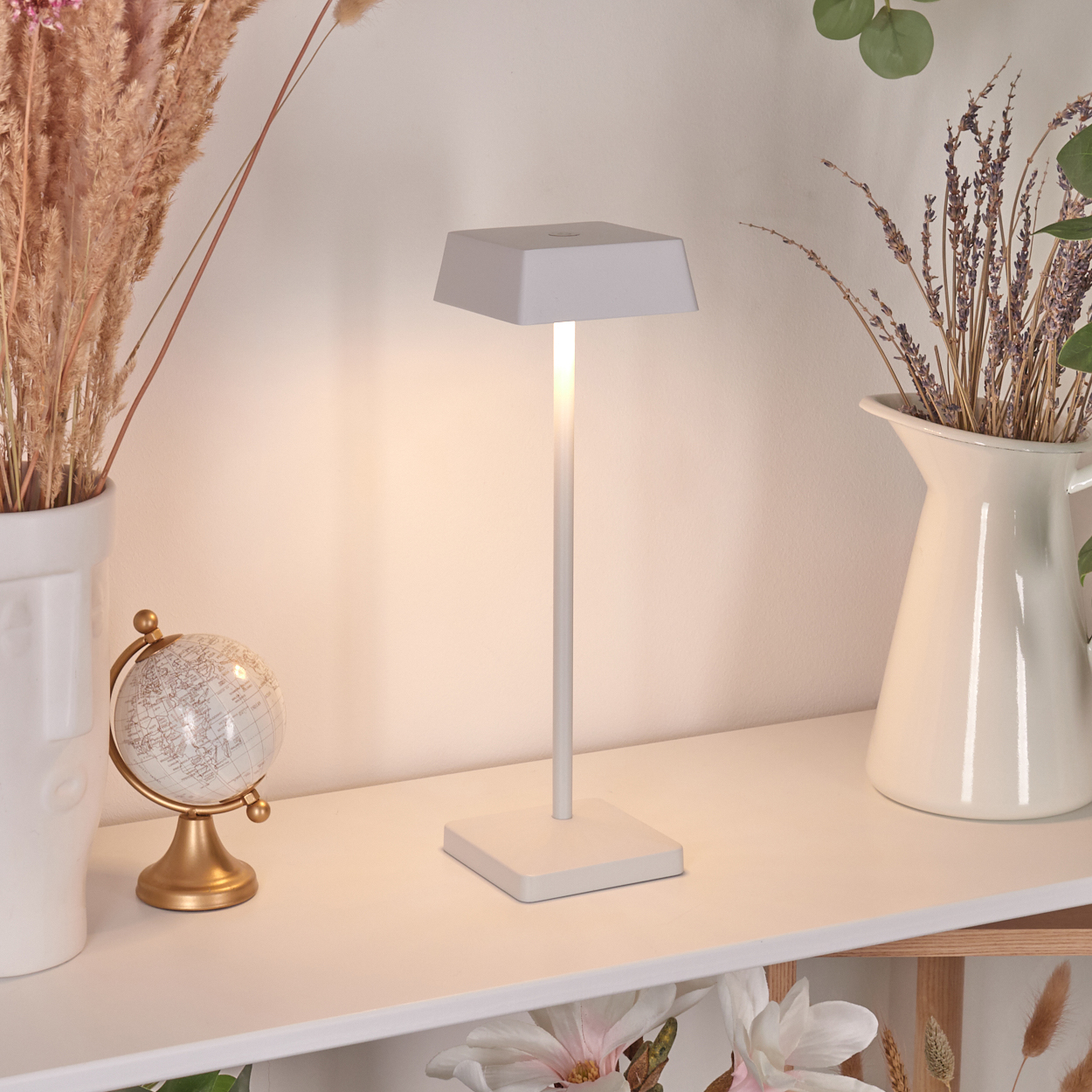 Eine weiße Tischlampe steht auf einem Regal neben Trockenblumen und einer Vase.