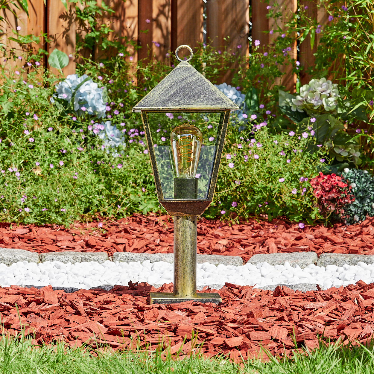 Ein goldener Gartenlaternenpfahl mit Glühbirne steht auf rotem Mulch und weißen Steinen.