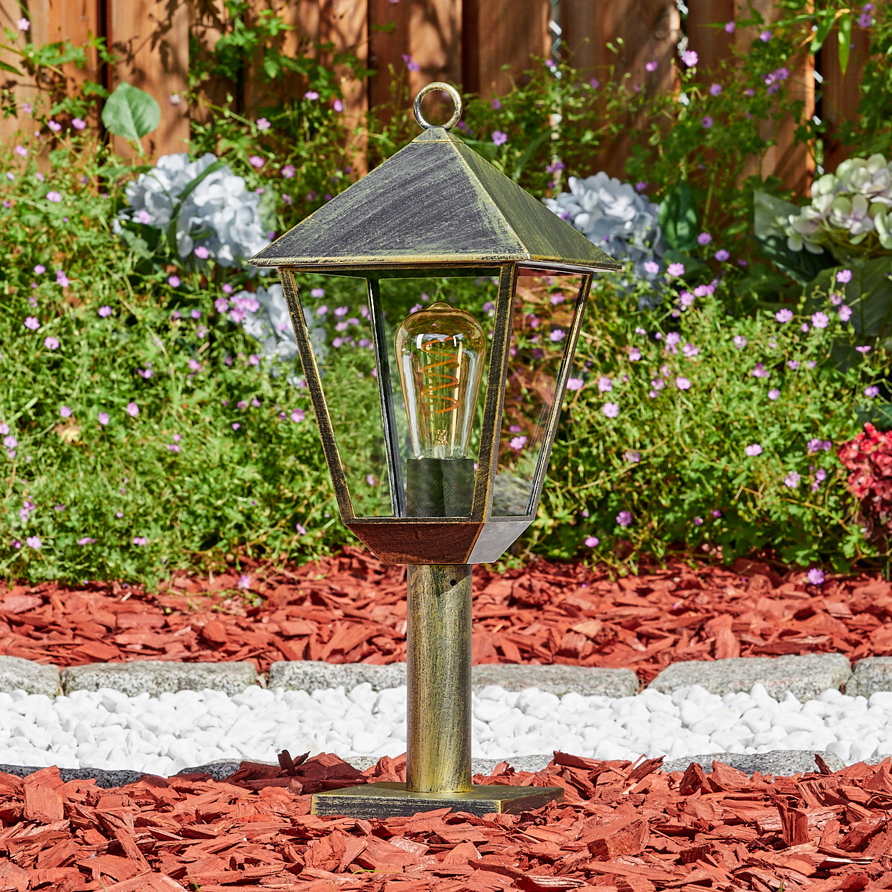 Eine goldene Gartenlaterne mit einer Glühbirne steht vor einem Holzzaun und Blumen.