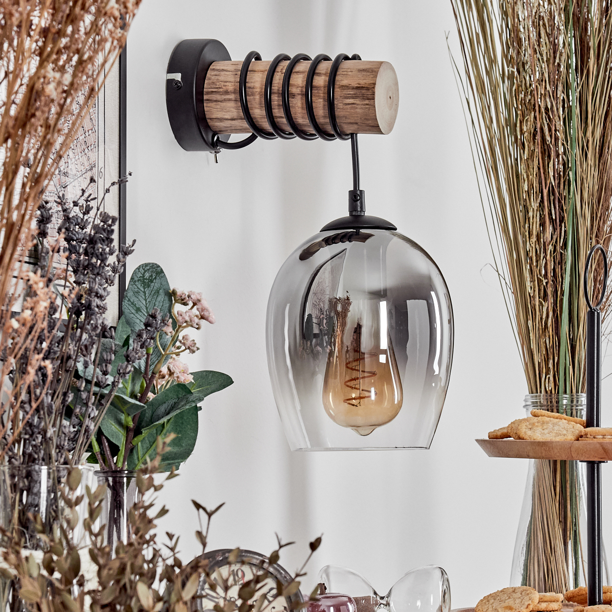 Eine Wandleuchte mit Holz, Metall und rauchigem Glaslampenschirm, daneben Trockenblumen.
