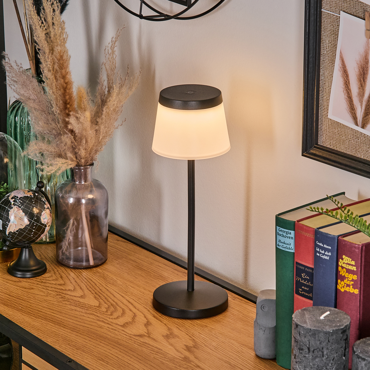 Eine schwarze Tischlampe leuchtet auf einem Holzregal neben Büchern und Dekoration.