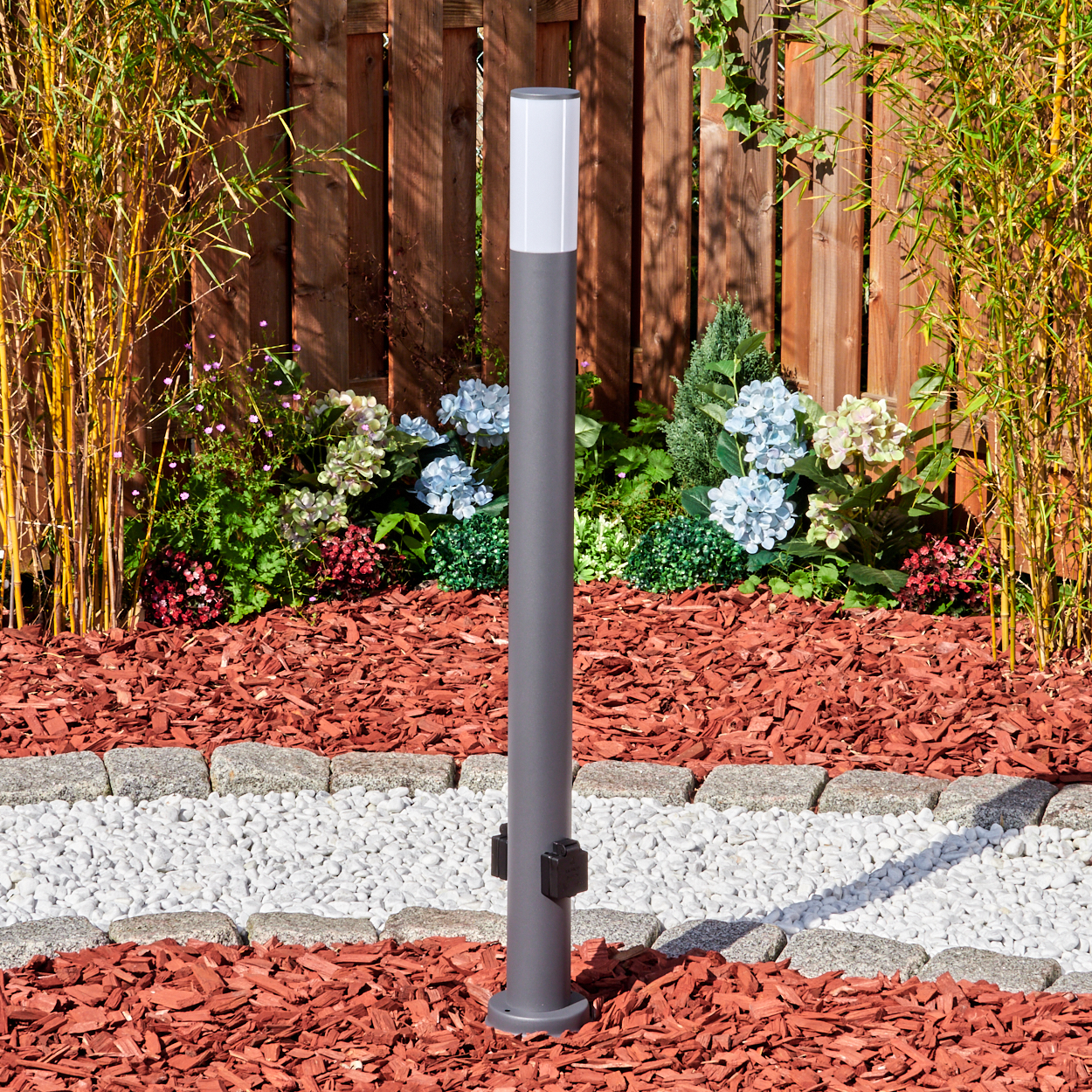 Ein grauer Außenposten mit weißem Lichtkopf steht in einem Garten mit rotem Mulch.