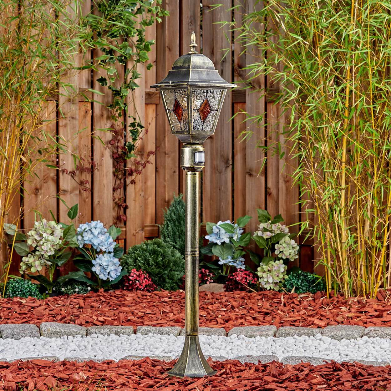 Ein antiker Gartenlichtmast mit Glasdetails steht vor einem Holzzaun und Bambus.