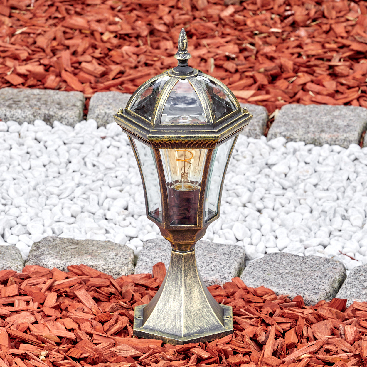 Ein bronzener Gartenpfosten mit Glaslaterne steht auf rotem Rindenmulch und weißen Kieselsteinen.