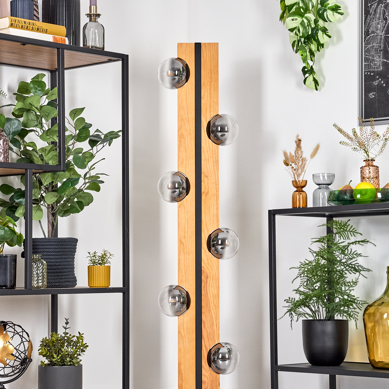 Ein vertikaler Holzsockel mit sechs Glaskugelleuchten steht neben Regalen mit Pflanzen und Deko.