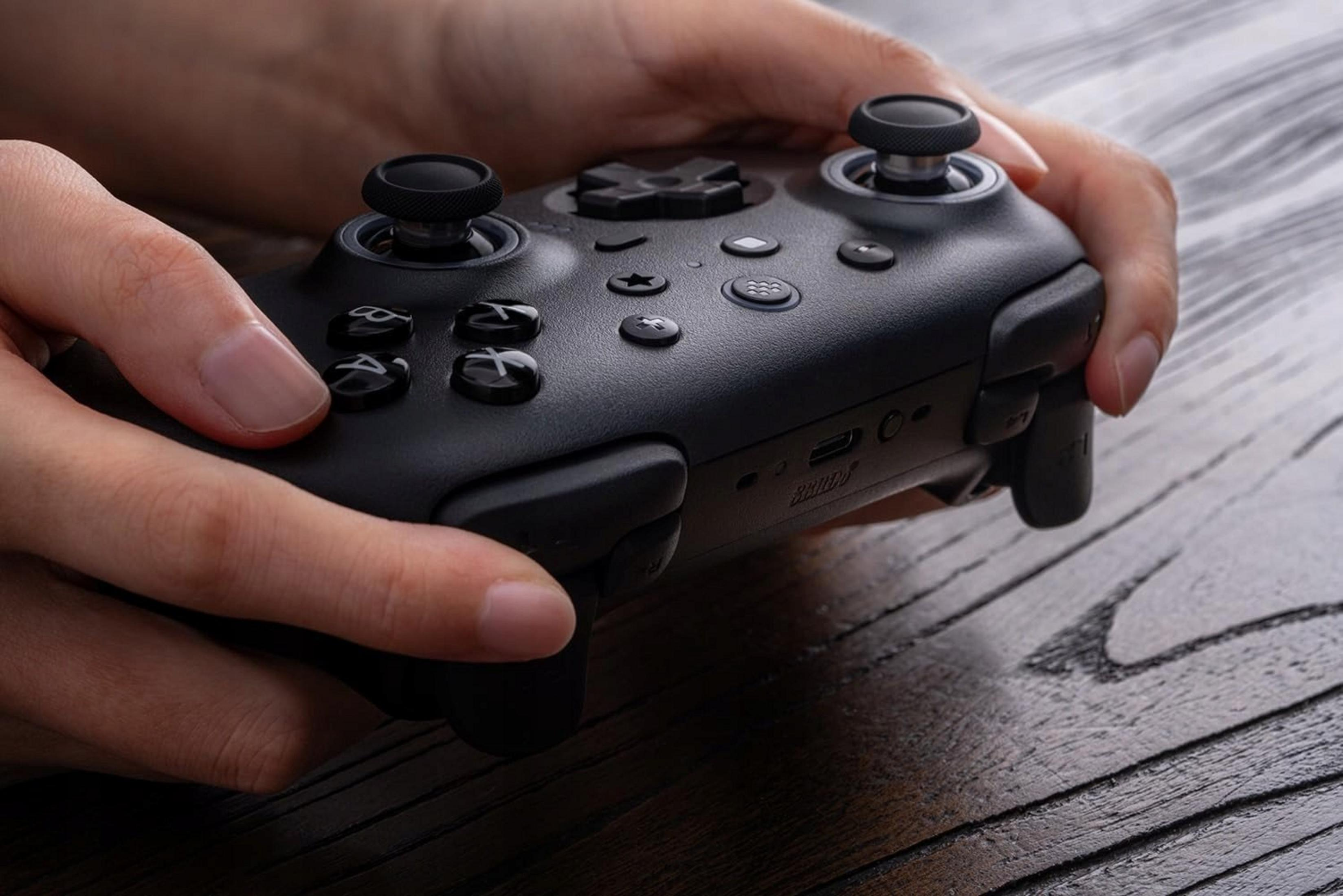 Hände halten einen schwarzen Gamecontroller auf einem Holztisch.