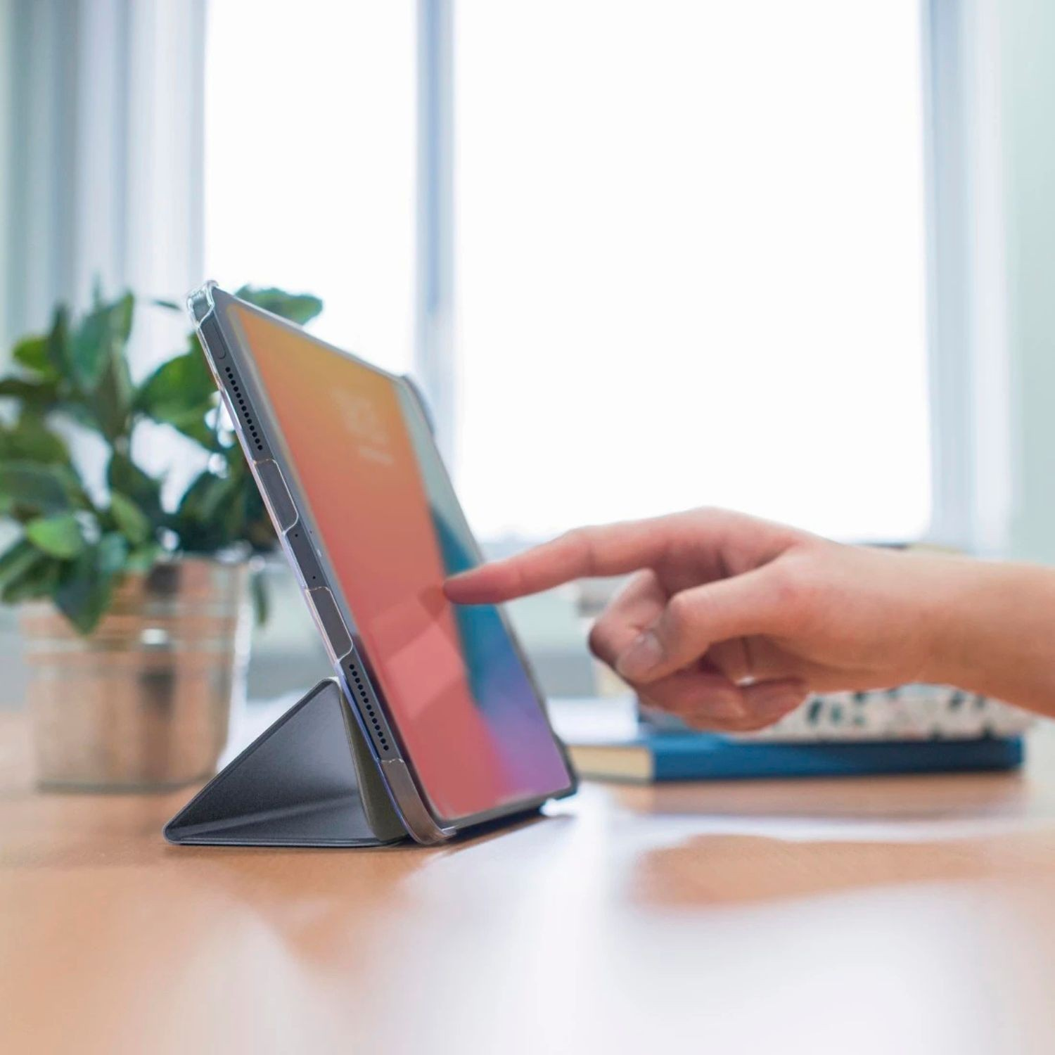 Dłoń dotyka tabletu postawionego na stojaku. W pobliżu roślina i książki.