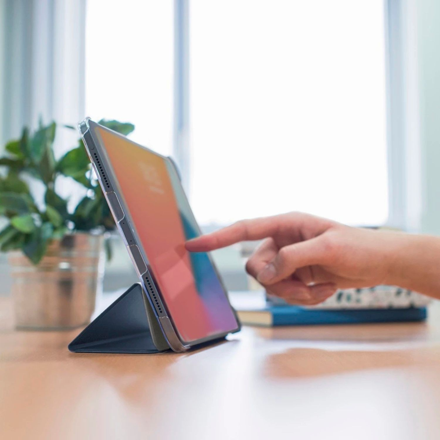 Dłoń dotyka ekranu tabletu na drewnianym biurku obok rośliny.