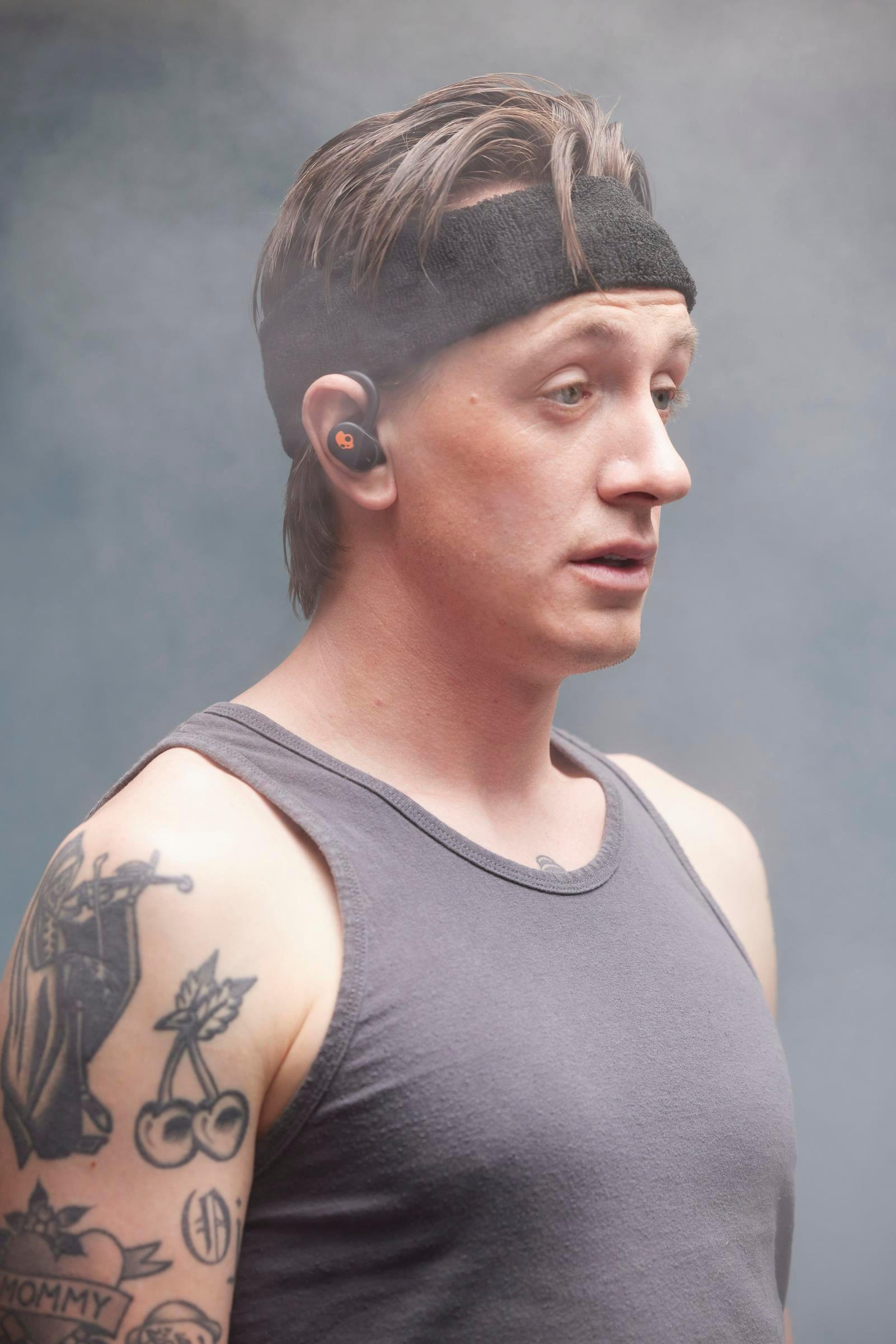 Mężczyzna w opasce i słuchawkach w profilu, szary podkoszulek, widoczne tatuaże.