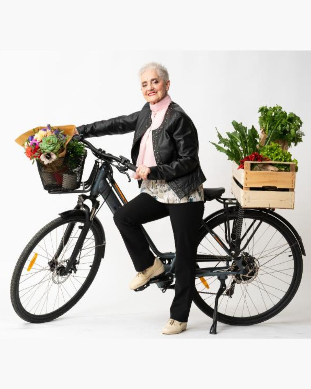 Donna anziana su bicicletta con cesto di fiori e cassa di verdure.