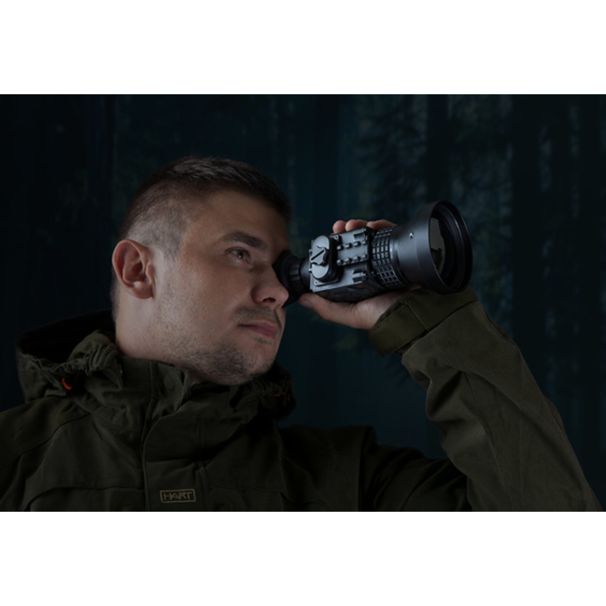 Ein Mann in einem grünen Anorak blickt durch ein Nachtsichtgerät in einem dunklen Wald.