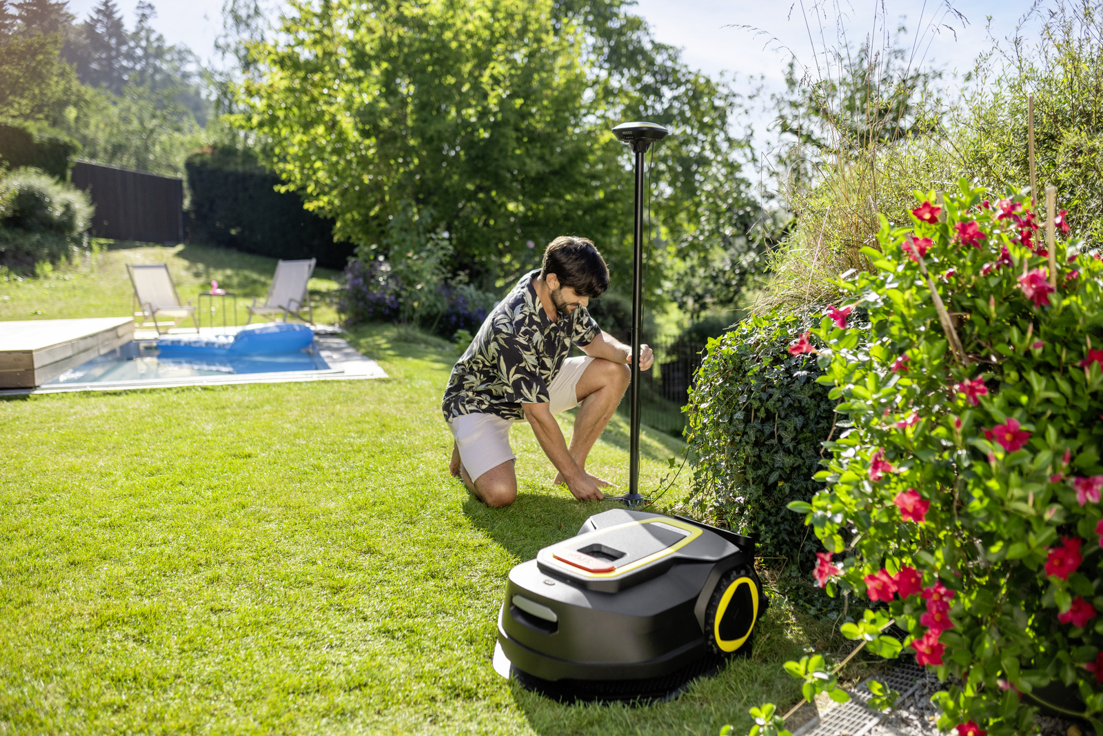Mann kniet neben Rasenroboter, Pool und Liegestühle im Hintergrund.