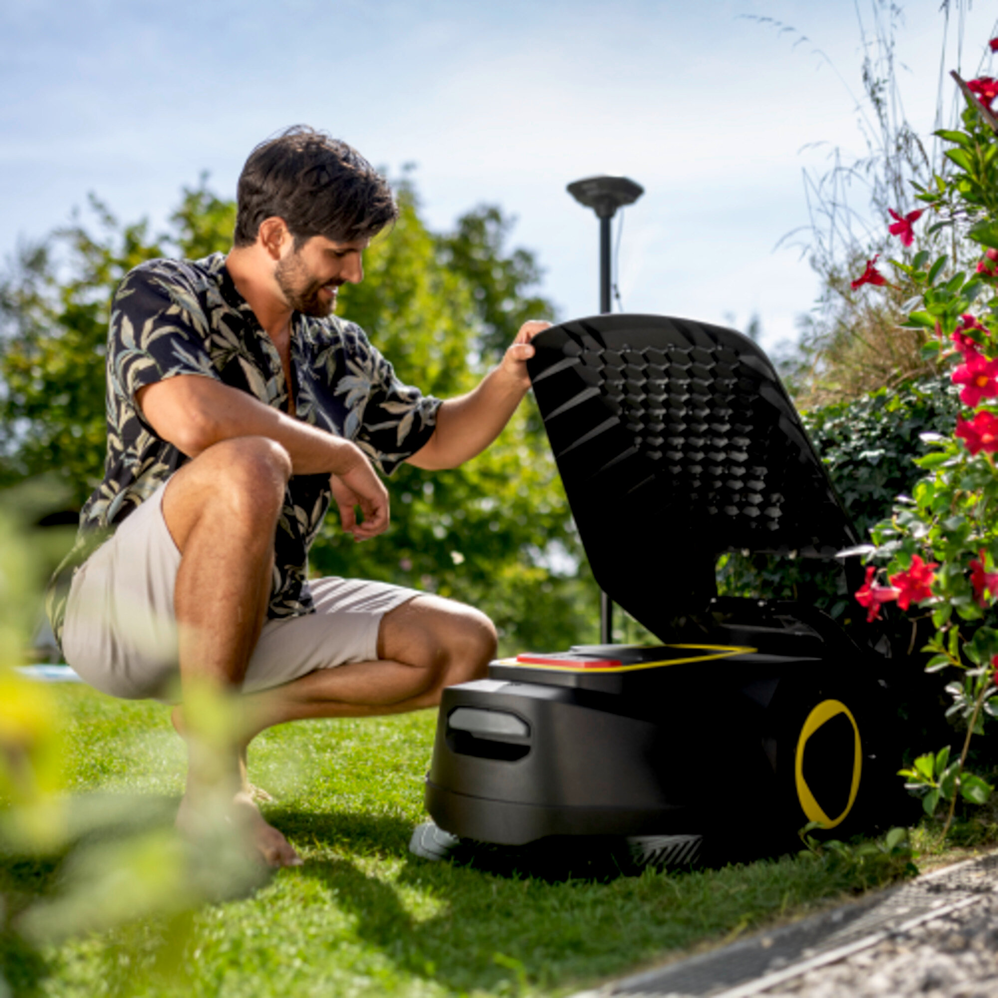 Mann im Blumenhemd kauert neben Rasenmähroboter mit offener Klappe, sonniger Garten.