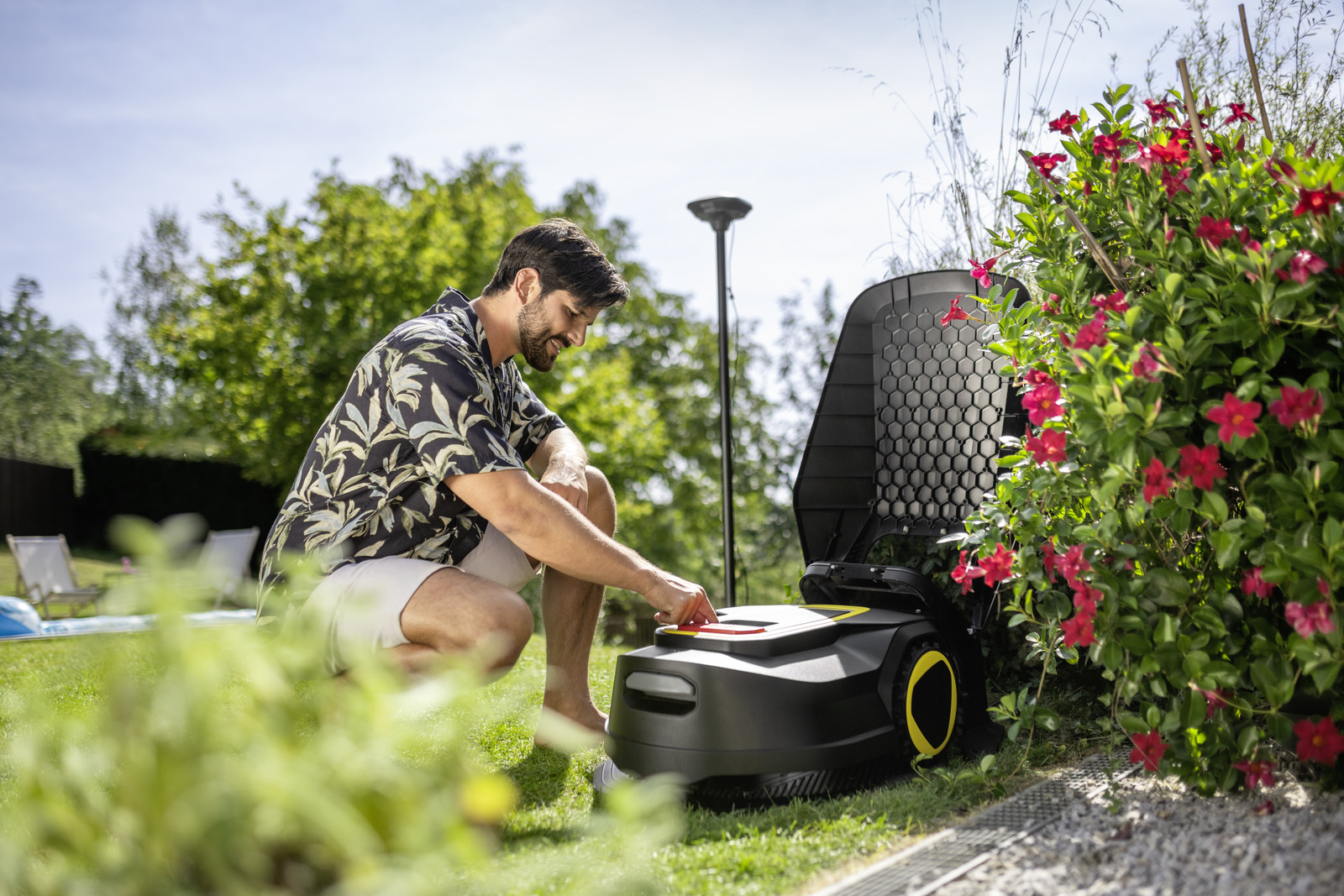 Mann im Blumenhemd stellt Rasenroboter bei roten Blumen und grünem Busch ein.