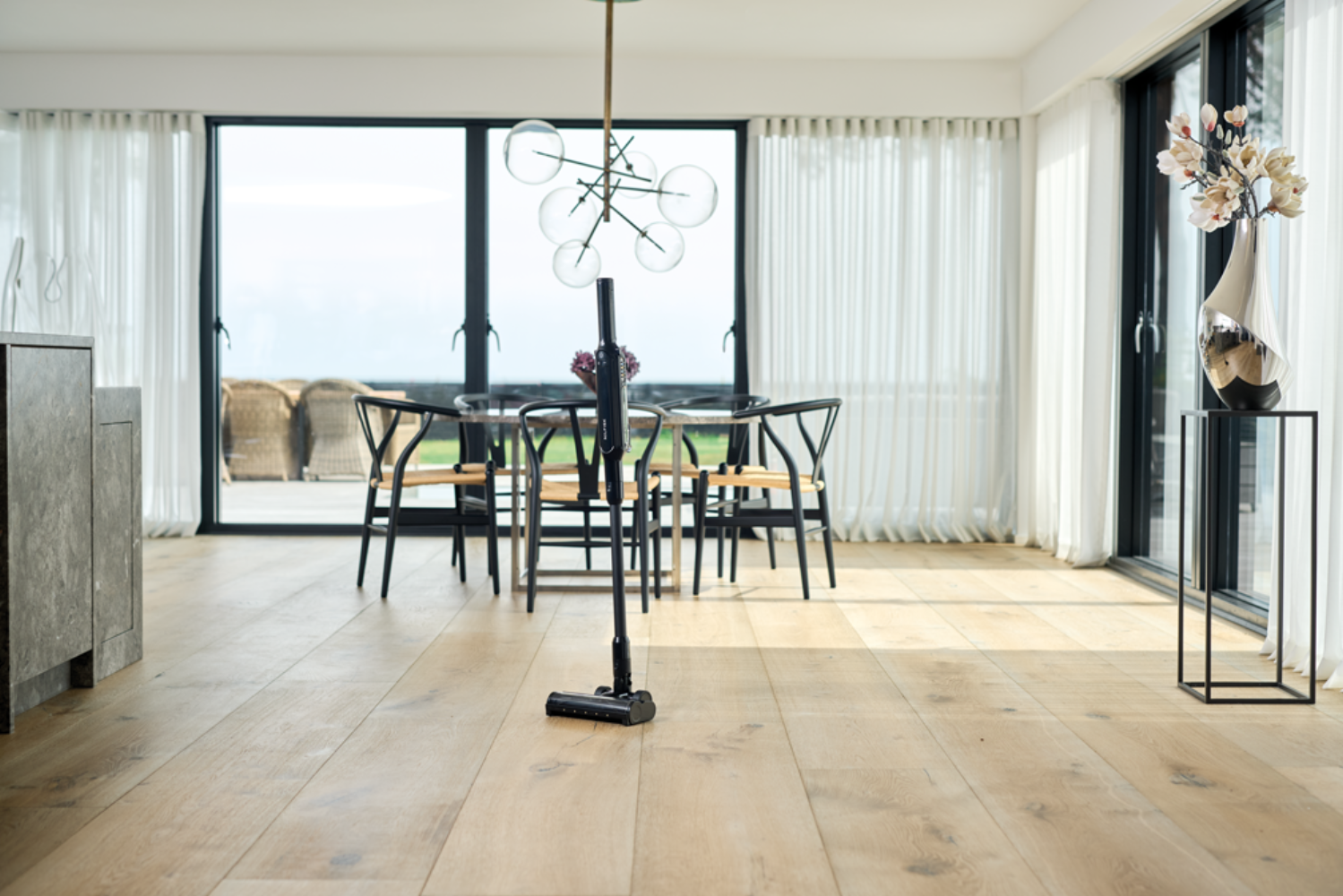 Una aspiradora inalámbrica sobre suelo de madera en un comedor con grandes ventanales.