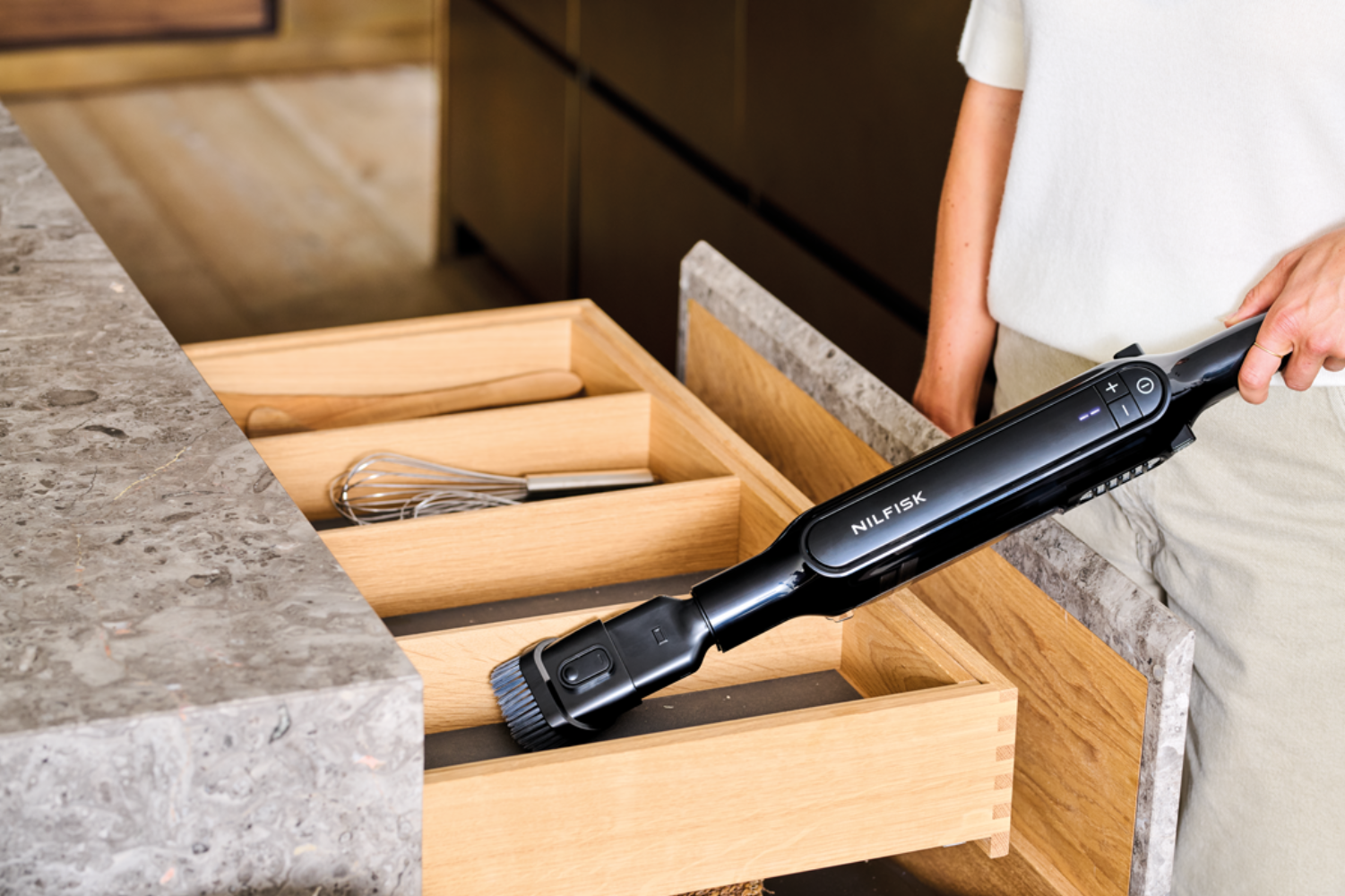 Una persona usa una aspiradora Nilfisk negra para limpiar un cajón de cocina de madera abierto.