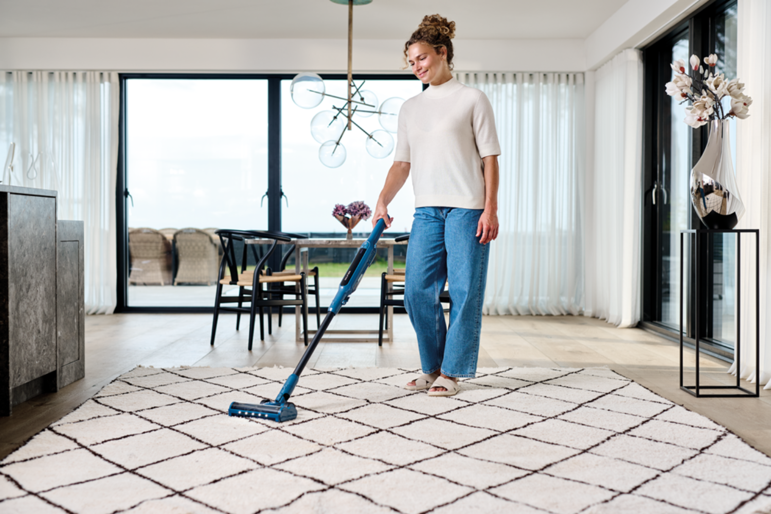 Una mujer aspira una alfombra en una habitación luminosa con grandes ventanales.