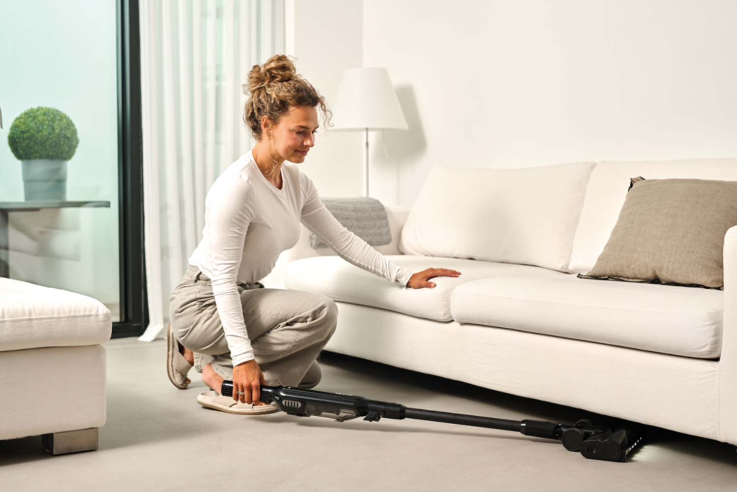 Eine Frau saugt unter einem weißen Sofa mit einem Stabstaubsauger.