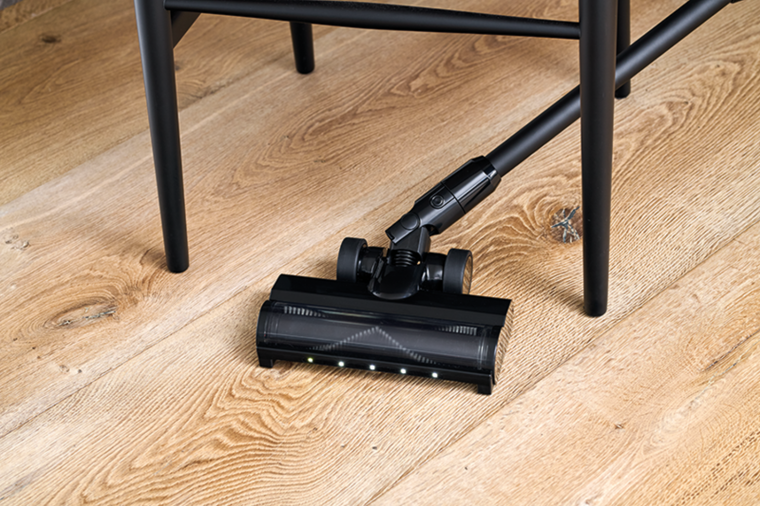 Cabezal de aspiradora negro con LEDs sobre suelo de madera, junto a patas de silla negras.