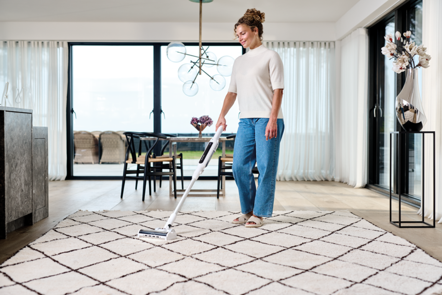 Una mujer aspira una alfombra en una sala moderna con grandes ventanales.