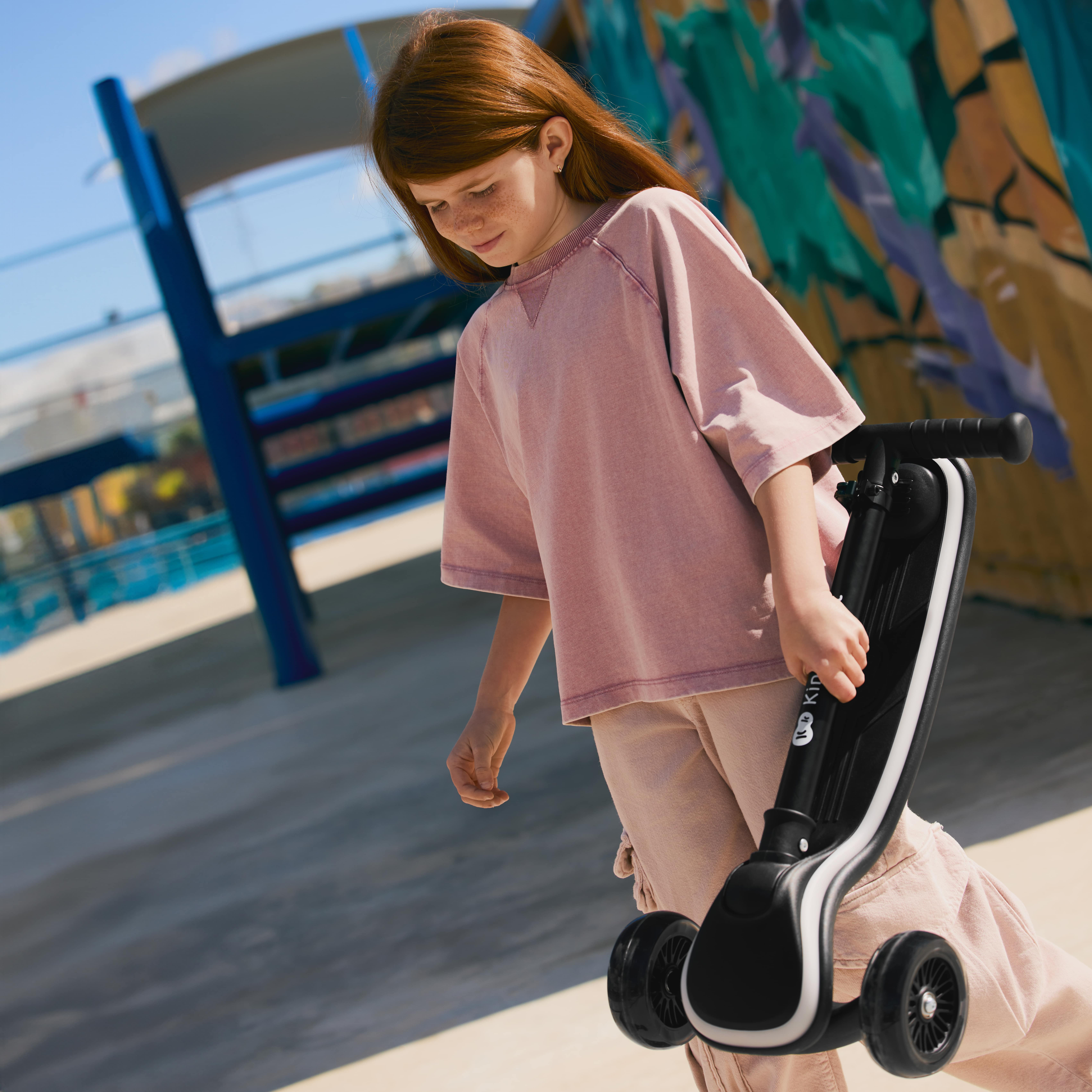 Ein Mädchen mit roten Haaren trägt ein rosa T-Shirt und hält einen schwarzen Roller.